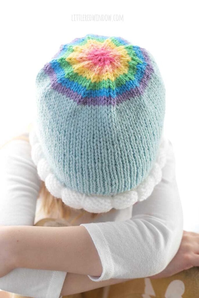 top view of blond girl wearing light blue knit hat with bubble knit white cloud brim and a rainbow decrease section on the top in front of a white background to show the rainbow circle on top of the hat