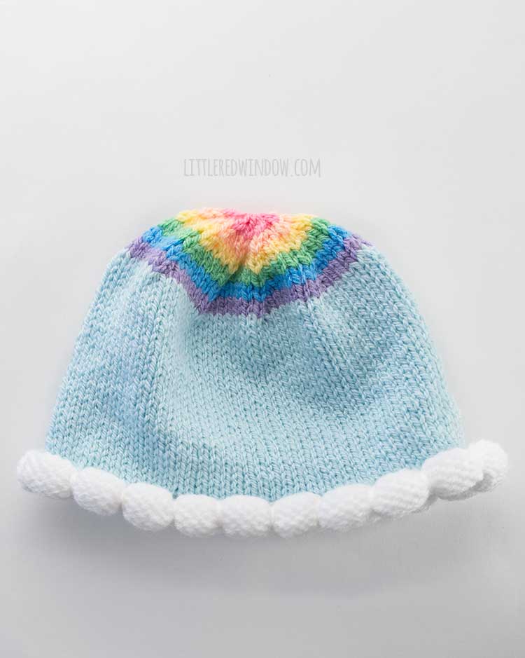 flay lay view of a light blue knit hat with puffy cloud bubble bim and round rainbow on top on a white tabletop