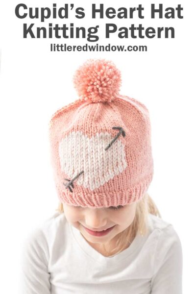 girl in white shirt looking down and wearing a light pink knit hat with large pink pom pom on top and a white heart with a charcoal gray arrow through it on the front of the hat