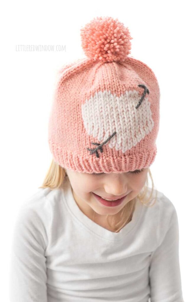 girl in white shirt looking down and laughing and wearing a light pink knit hat with large pink pom pom on top and a white heart with a charcoal gray arrow through it on the front of the hat all in front of a white background