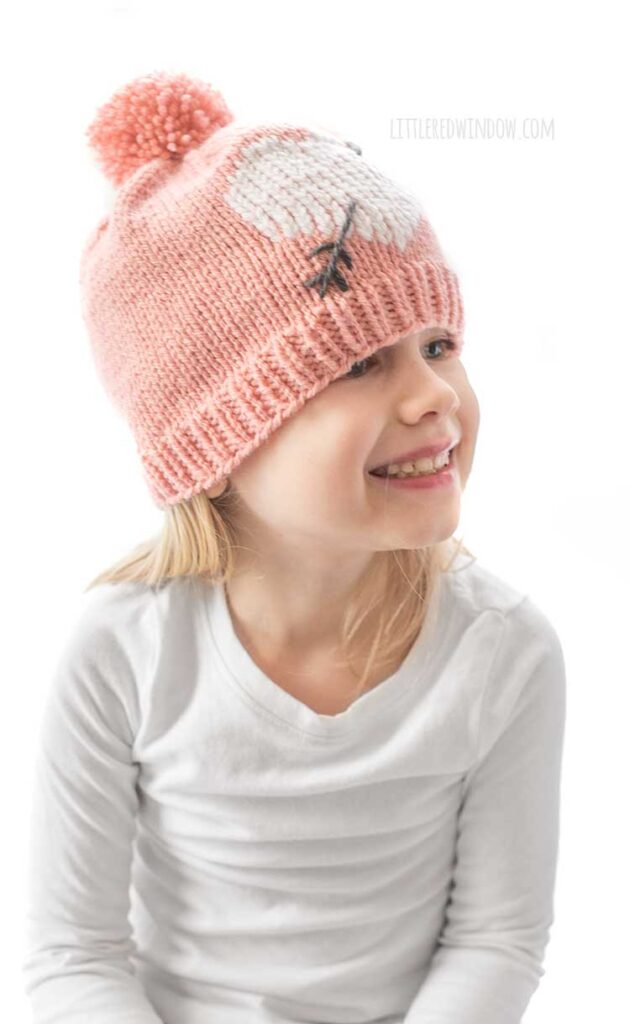 girl in white shirt looking up and to the right and laughing and wearing a light pink knit hat with large pink pom pom on top and a white heart with a charcoal gray arrow through it on the front of the hat all in front of a white background