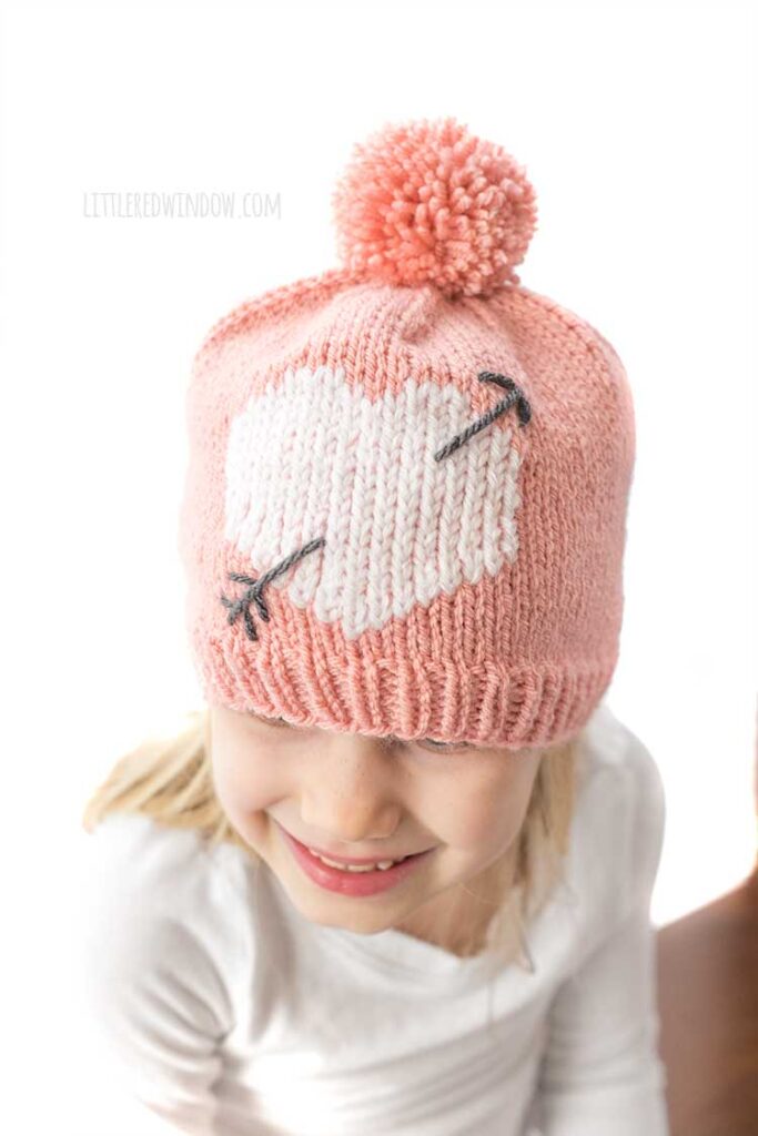 close up view from slightly above of girl in white shirt looking down and wearing a light pink knit hat with large pink pom pom on top and a white heart with a charcoal gray arrow through it on the front of the hat all in front of a white background