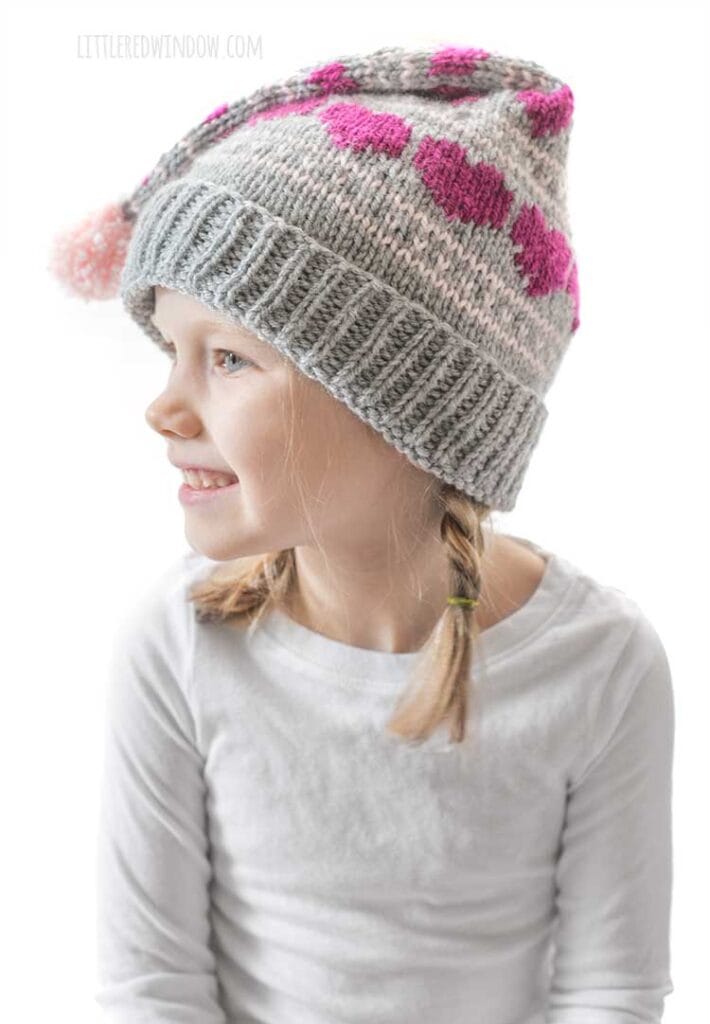 smiling girl in white shirt wearing a gray knit stocking cap with magenta hearts knit on it looking off to the left in front of a white background