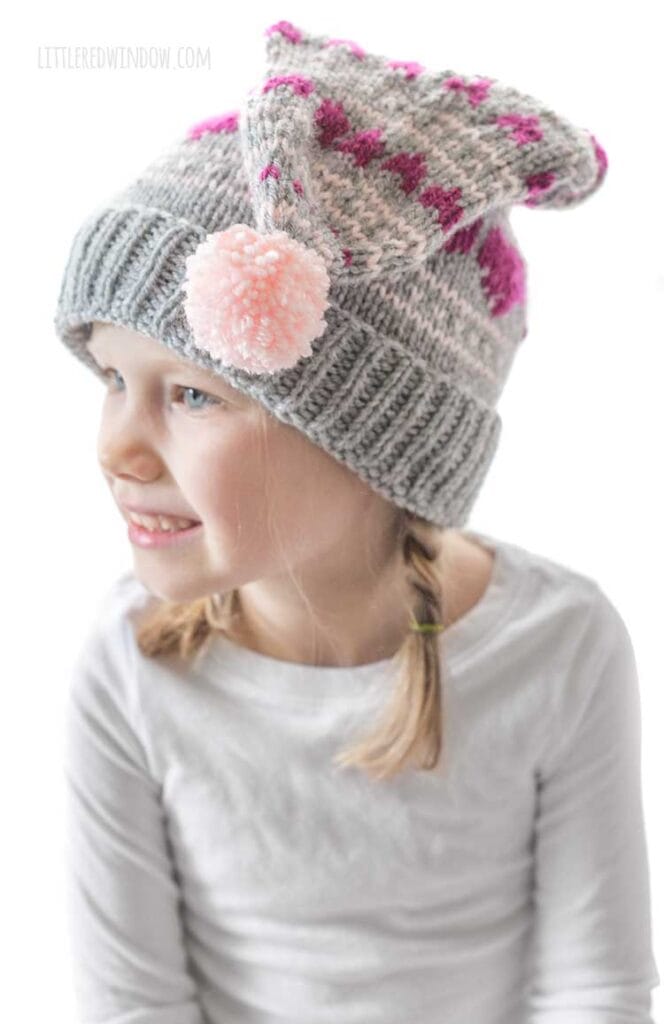 smiling girl in white shirt wearing a gray knit stocking cap with magenta hearts knit on it and a pink pom pom in front looking off to the left in front of a white background