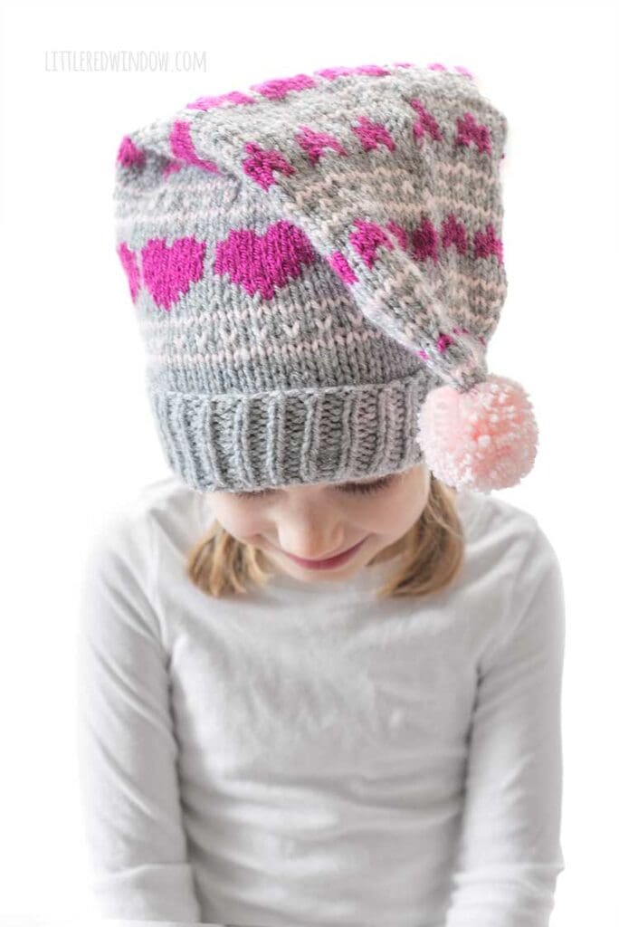 smiling girl in white shirt wearing a gray knit stocking cap with magenta hearts knit on it and a pink pom pom in front looking down in front of a white background