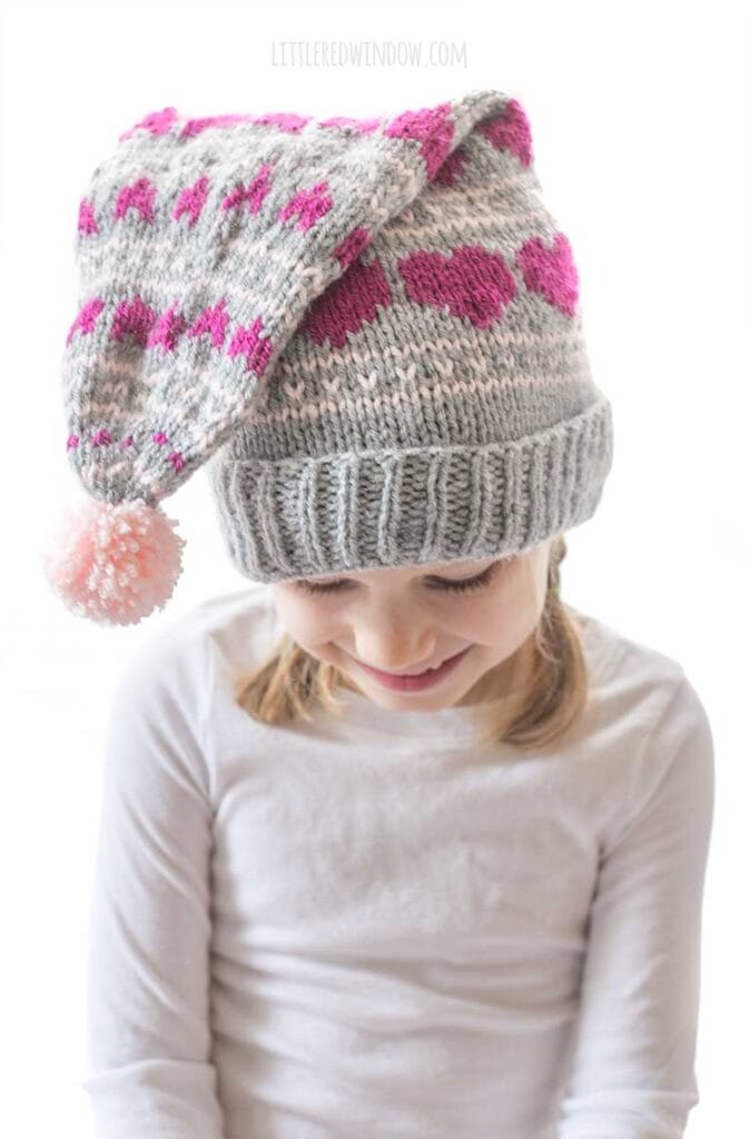 smiling girl in white shirt wearing a gray knit stocking cap with magenta hearts knit on it and a pink pom pom in front looking down in front of a white background