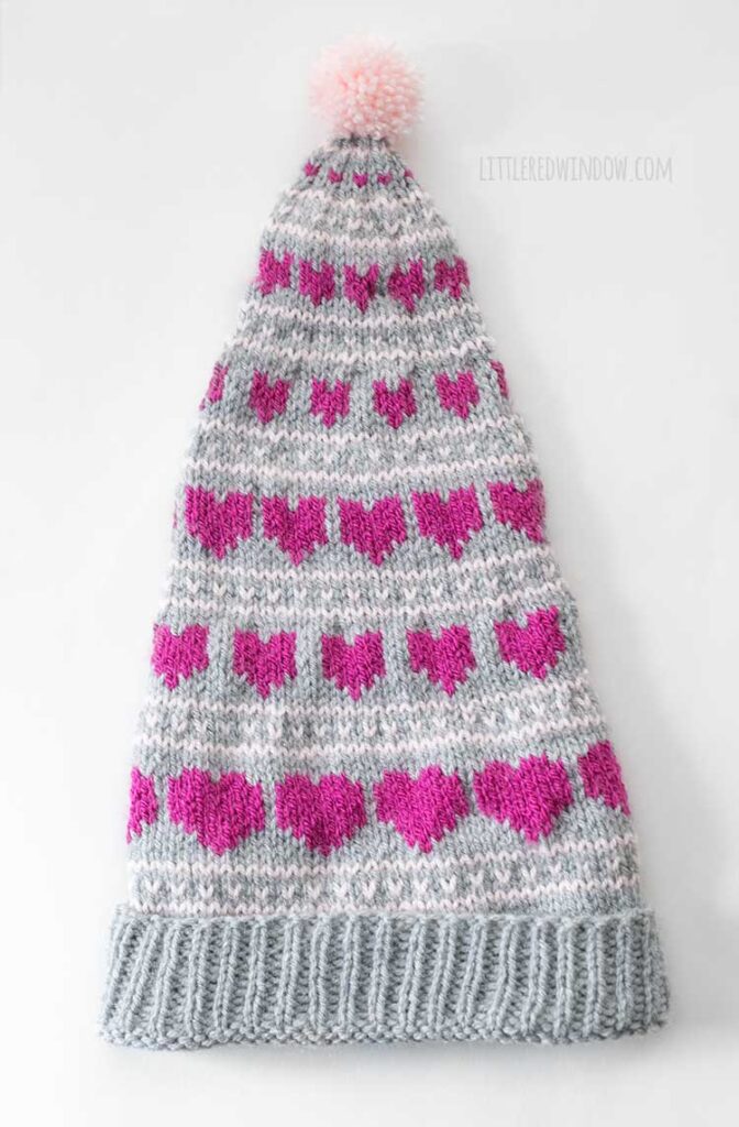 Flat lay image of a gray knit stocking cap on a white background with an alternating pattern of light pink lines and magenta hearts The hearts get small as they closer to the top of the hat