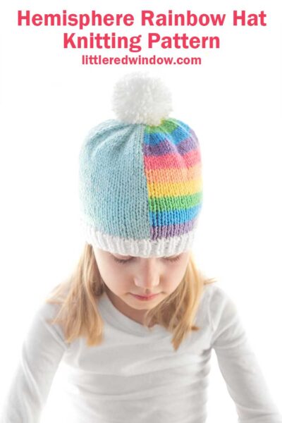 view of blond girl in white shirt looking down at her lap and wearing a knit hat that is half light blue and half pastel rainbow stripes and has a large white pom pom on top