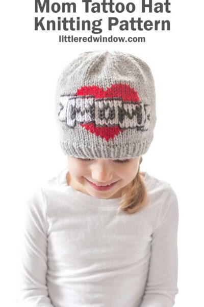girl in a white shirt in front of a white background looking down at her lap and wearing a gray knit beanie with the classic mom tattoo motif on the front a red heart with a white banner in front that reads MOM