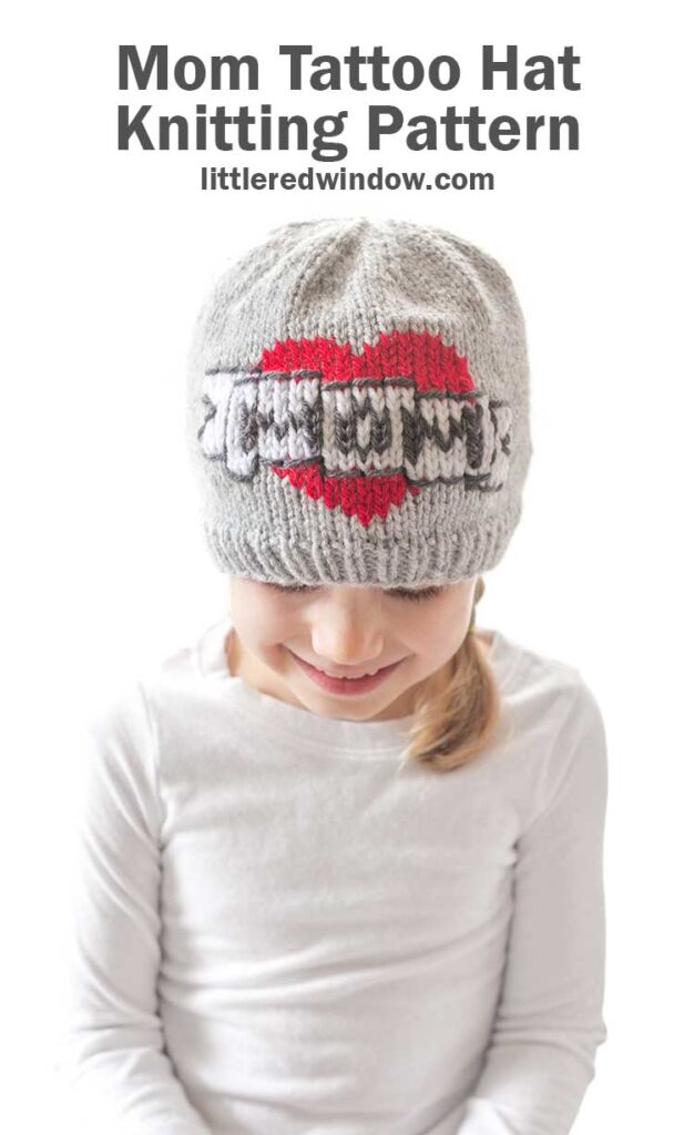 girl in a white shirt in front of a white background looking down at her lap and wearing a gray knit beanie with the classic mom tattoo motif on the front a red heart with a white banner in front that reads MOM