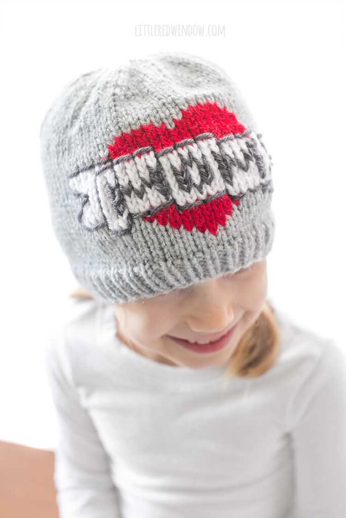 girl in a white shirt in front of a white background looking down off to the right and wearing a gray knit beanie with the classic mom tattoo motif on the front a red heart with a white banner in front that reads MOM