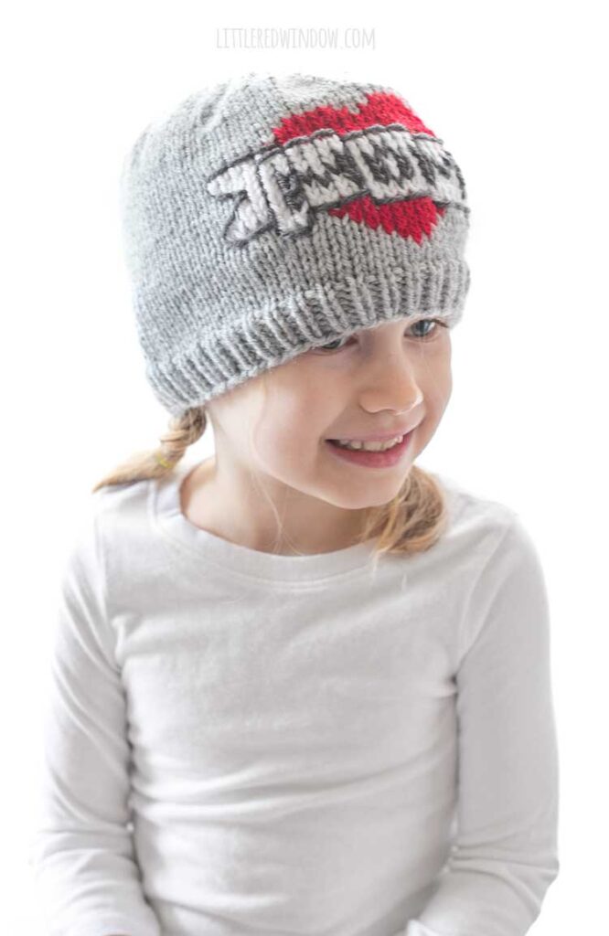 girl in a white shirt in front of a white background looking slightly right of the cameraand wearing a gray knit beanie with the classic mom tattoo motif on the front a red heart with a white banner in front that reads MOM