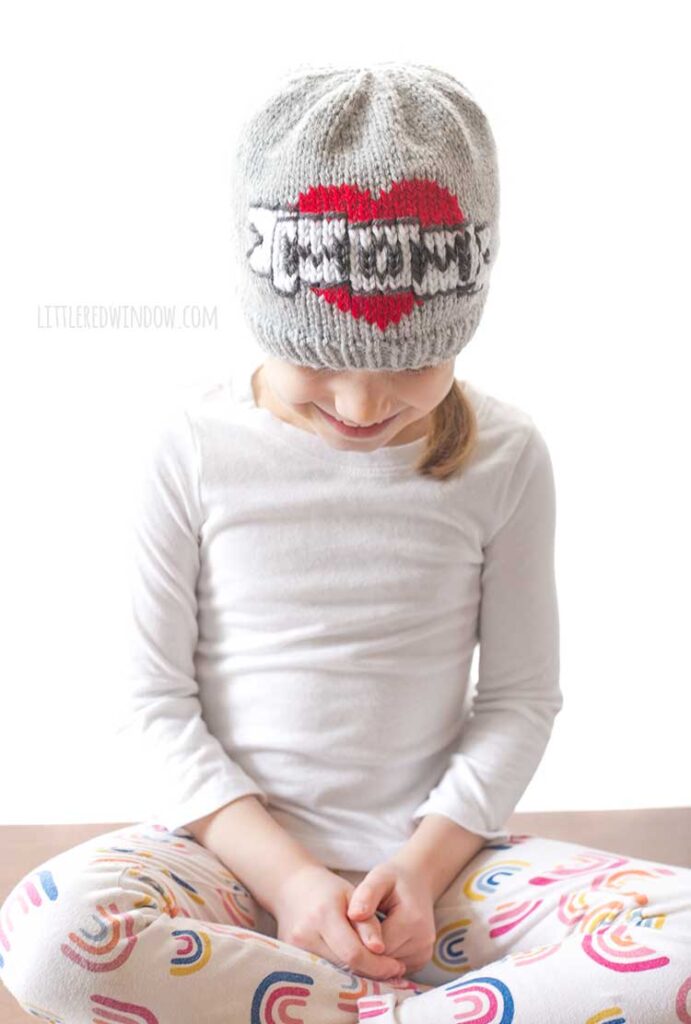 farther view of girl in a white shirt sitting cross legged in front of a white background looking down at her lap and wearing a gray knit beanie with the classic mom tattoo motif on the front a red heart with a white banner in front that reads MOM