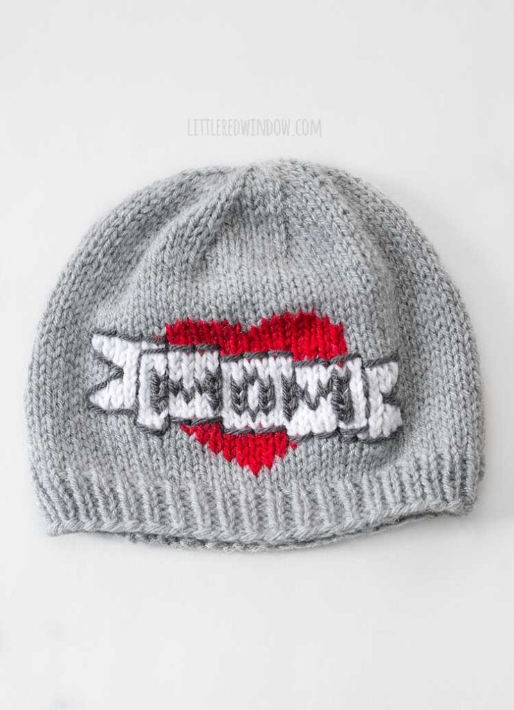 flat lay image of a gray knit hat with the classic mom tattoo motif on the front of a large red heart with a white banner in front of it that says MOM