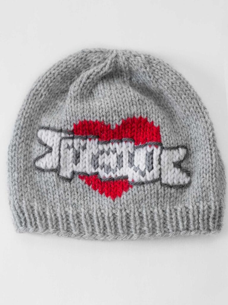 flat lay image of a gray knit hat with the classic mom tattoo motif on the front of a large red heart with a white banner in front of it that says YOU