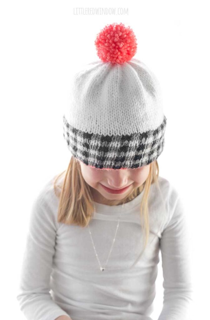girl in white shirt wearing a white knit hat the black and white plaid brim and pink pom pom on top looking down