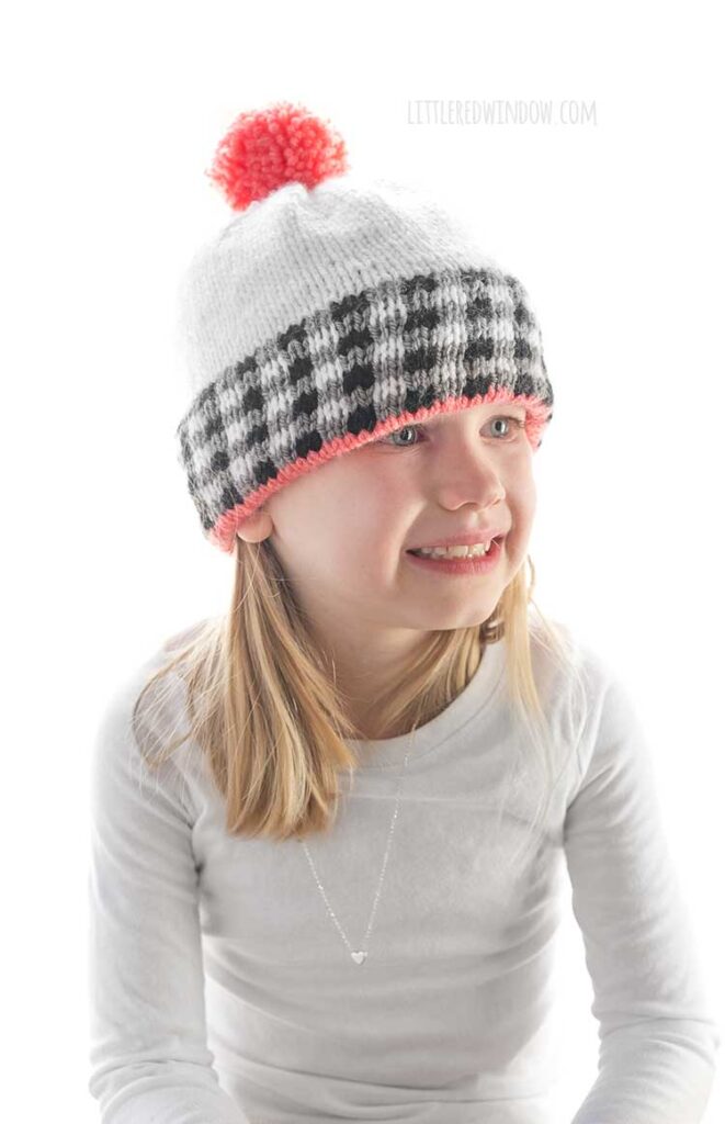 girl in white shirt wearing a white knit hat the black and white plaid brim and pink pom pom on top smiling and looking off to the right