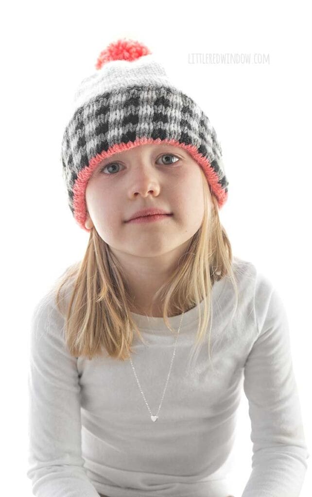 girl in white shirt wearing a white knit hat the black and white plaid brim and pink pom pom on top looking at the camera