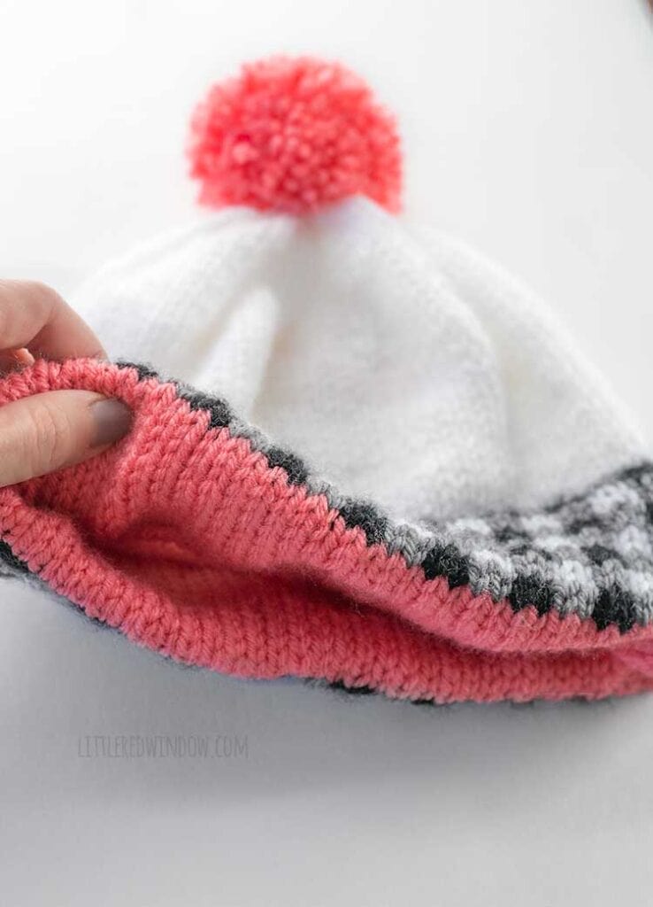 a hand lifting up the edge of the plaid double brim hat to show the double thick pink stockinette stitch lining inside as it is lying on a white tabletop