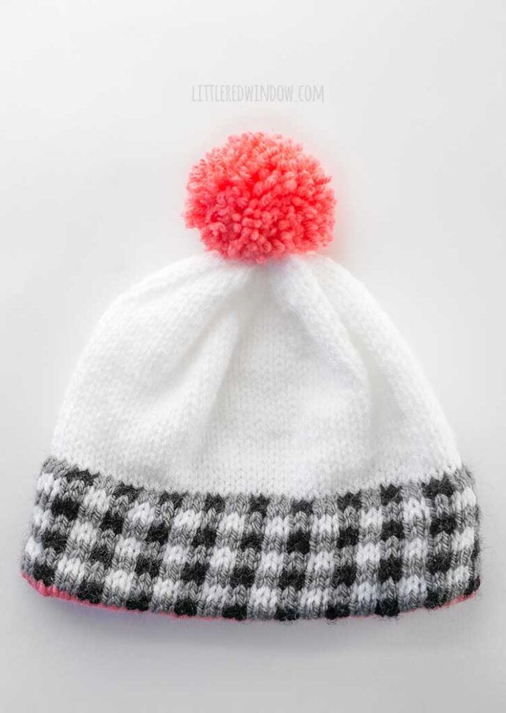 white knit hat with black and white plaid brim and pink pom pom on top lying flat on a white tabletop