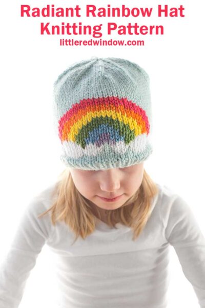 girl in white shirt wearing a light blue knit hat with a six color rainbow on the front center looking down at their lap