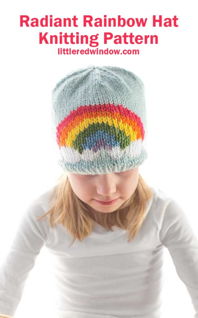 girl in white shirt wearing a light blue knit hat with a six color rainbow on the front center looking down at their lap