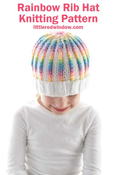 image showing a girl in a white shirt wearing a knit hat with a pattern of white and rainbow striped ribbing and a fully white folded brim. She is looking down at her lap in front of a white background