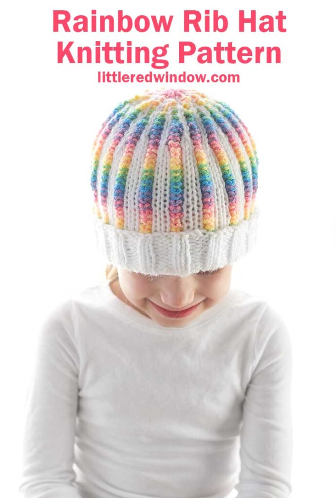 image showing a girl in a white shirt wearing a knit hat with a pattern of white and rainbow striped ribbing and a fully white folded brim. She is looking down at her lap in front of a white background
