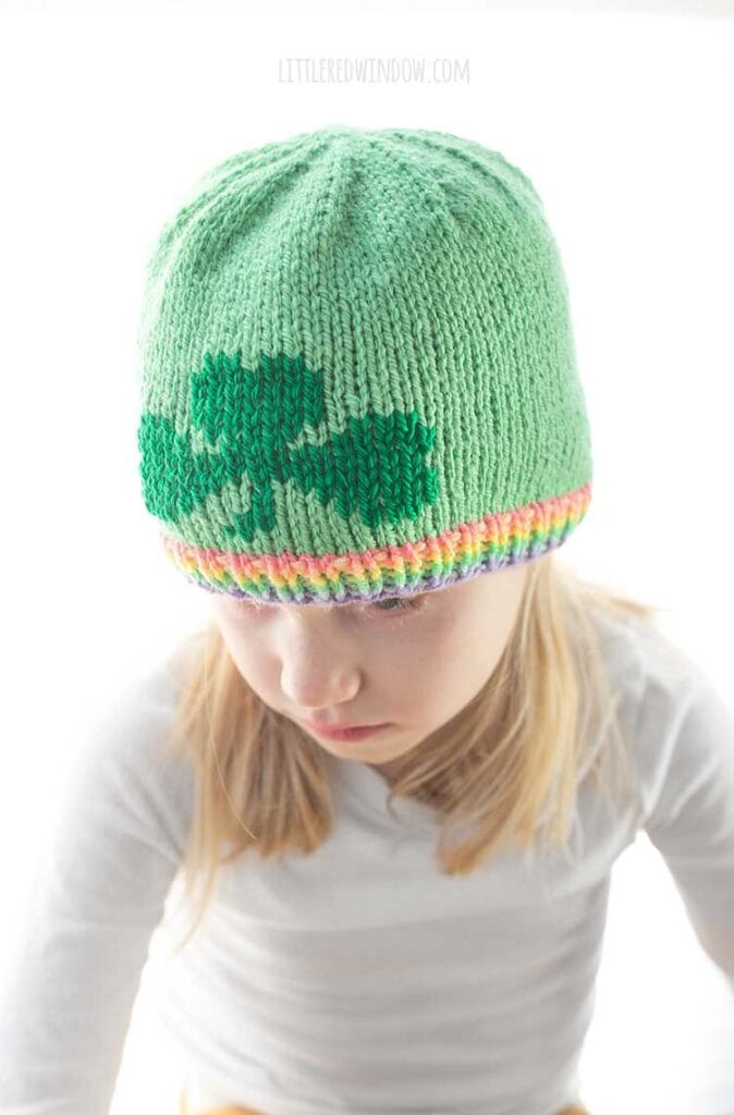 blond girl in white long sleeved shirt wearing a light green knit hat with a pastel rainbow ribbed brim and a darker green shamrock on the front looking down and to the left