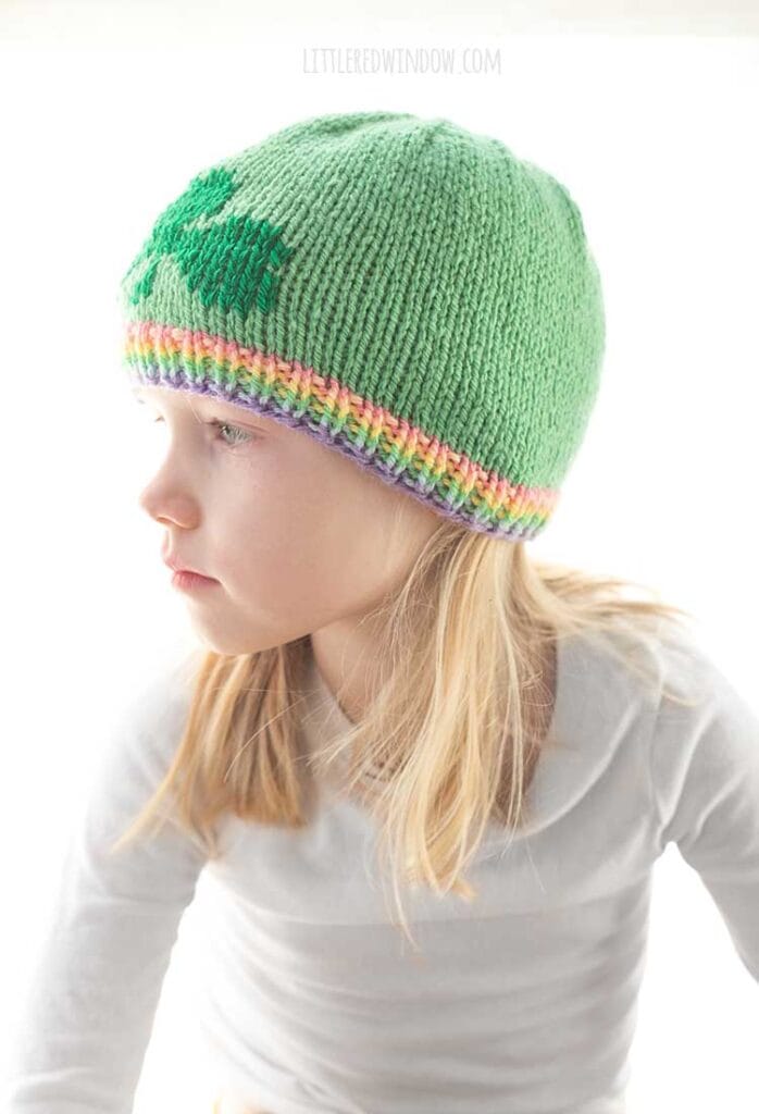 blond girl in white long sleeved shirt wearing a light green knit hat with a pastel rainbow ribbed brim and a darker green shamrock on the front looking off to the left