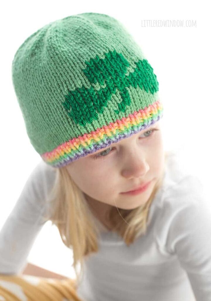 view from slightly above showing blond girl in white long sleeved shirt wearing a light green knit hat with a pastel rainbow ribbed brim and a darker green shamrock on the front looking off to the right