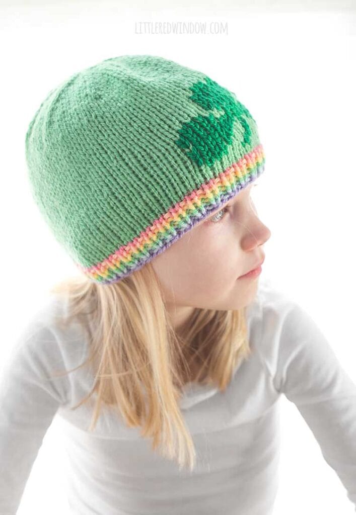 blond girl in white long sleeved shirt wearing a light green knit hat with a pastel rainbow ribbed brim and a darker green shamrock on the front looking off to the right