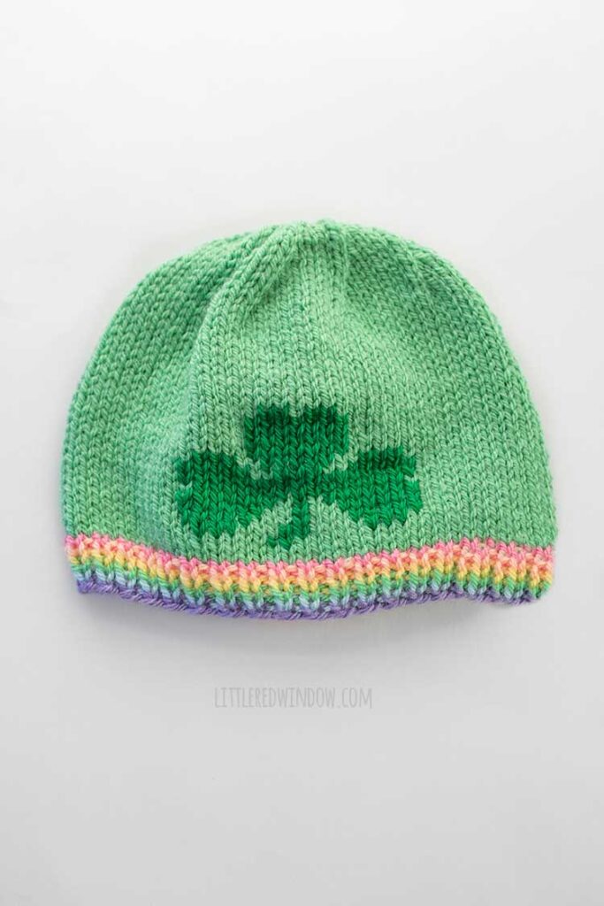 flat lay view of a light green knit hat with a pastel ribbed brim and a dark green three leaf shamrock shape on the front in duplicate stitch lying on a white tabletop