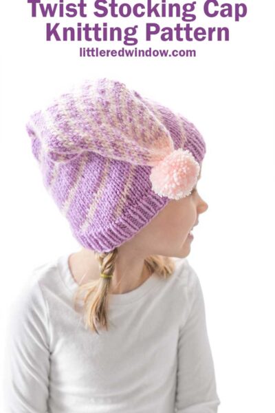 girl in white long sleeved shirt in front of a white background looking back over her left shoulder and wearing a light purple stocking cap style hat folded down toward the camera with a pattern of twisting light pink lines getting closer together at the top and ending in a light pink pom pom