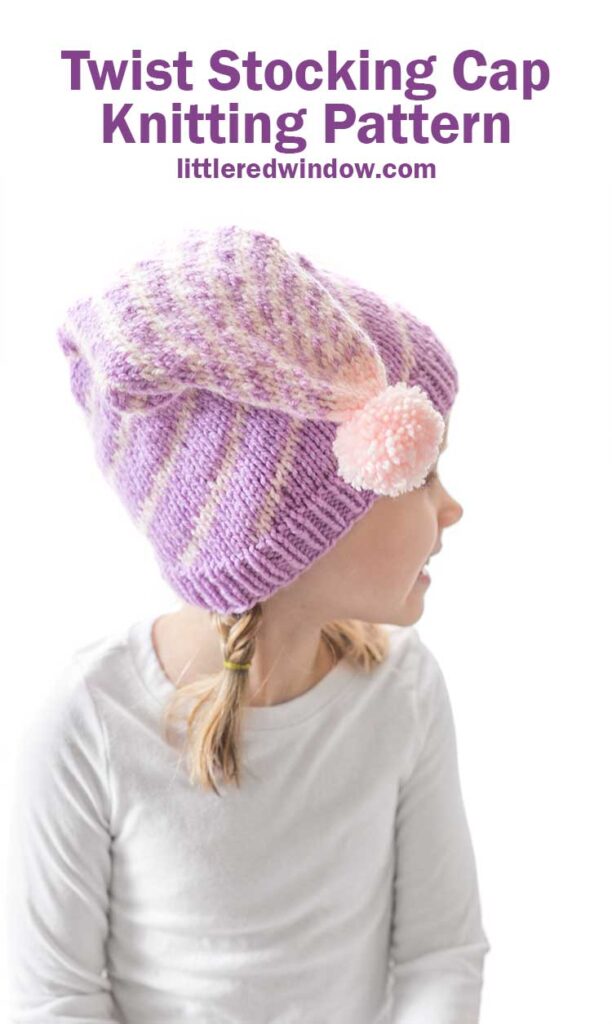 girl in white long sleeved shirt in front of a white background looking back over her left shoulder and wearing a light purple stocking cap style hat folded down toward the camera with a pattern of twisting light pink lines getting closer together at the top and ending in a light pink pom pom