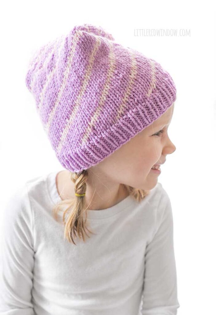 girl in white long sleeved shirt in front of a white background looking back over her left shoulder and wearing a light purple stocking cap style hat folded down away from the camera with a pattern of twisting light pink lines getting closer together at the top and ending in a light pink pom pom
