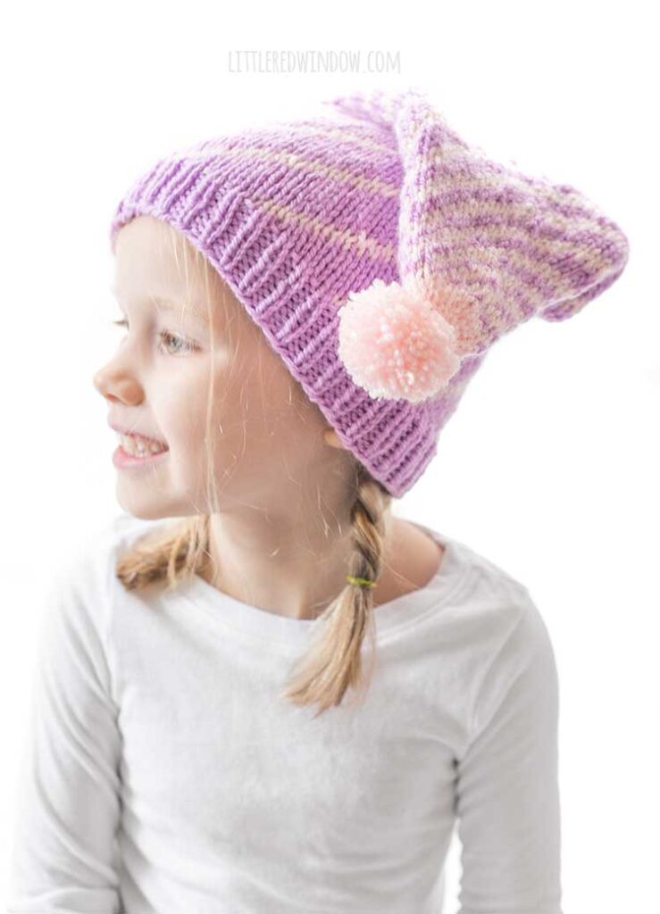 girl in white long sleeved shirt in front of a white background looking back off to the left and wearing a light purple stocking cap style hat folded down toward the camera with a pattern of twisting light pink lines getting closer together at the top and ending in a light pink pom pom