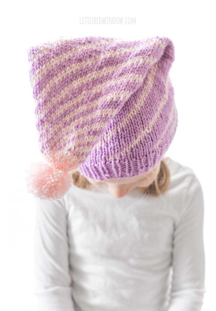 girl in white long sleeved shirt in front of a white background looking way down at her lap and wearing a light purple stocking cap style hat folded straight forward toward the camera with a pattern of twisting light pink lines getting closer together at the top and ending in a light pink pom pom