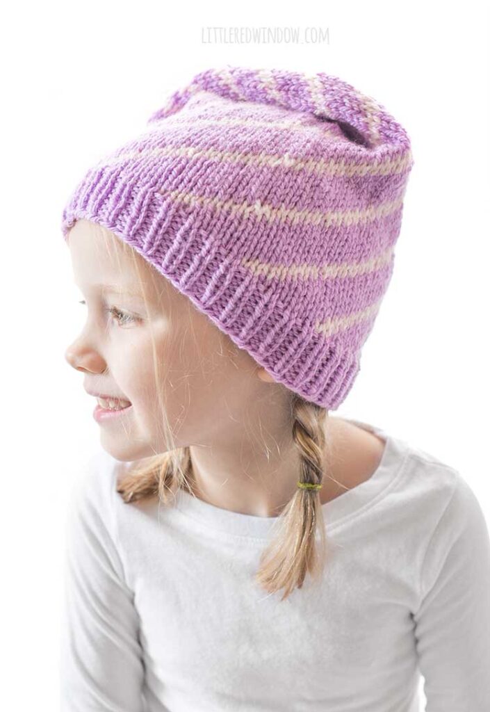 girl in white long sleeved shirt in front of a white background looking back off to the left and wearing a light purple stocking cap style hat folded down away from the camera with a pattern of twisting light pink lines getting closer together at the top and ending in a light pink pom pom