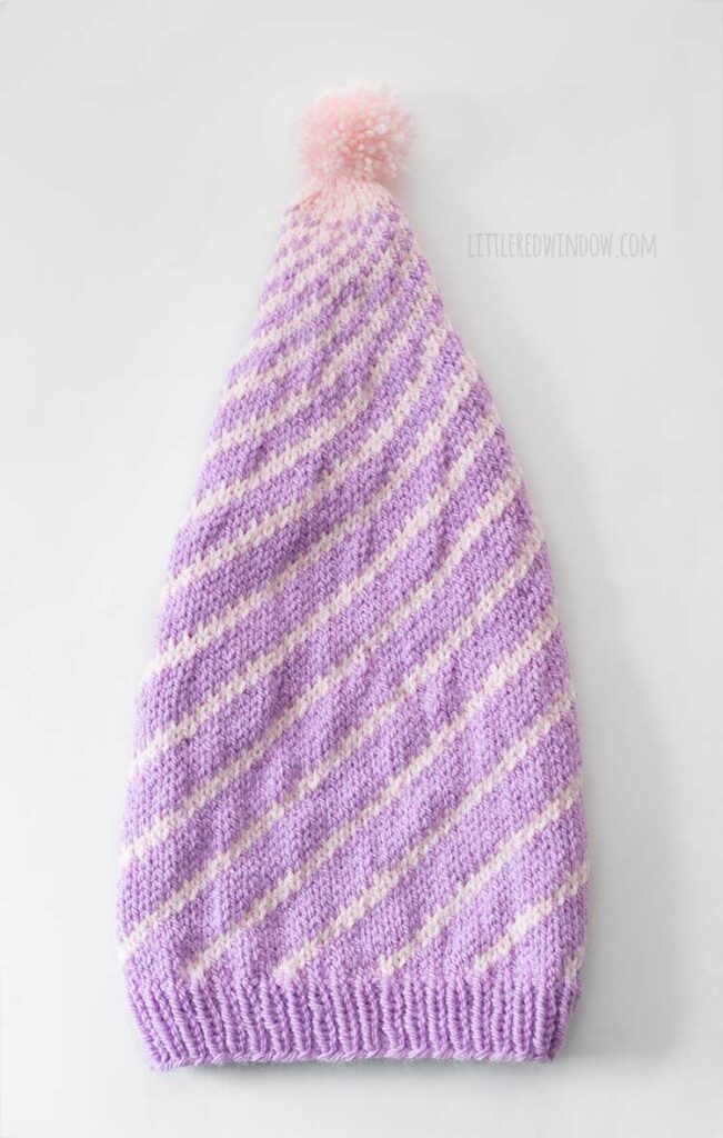 flat lay image of light purple knit stocking cap with pattern of light pink lines that twist around the hat getting closer together towards the top where this is a light pink pom pom