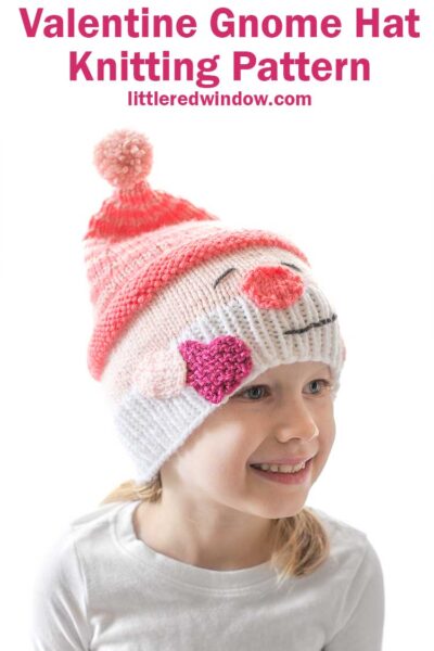 smiling girl in white shirt in front of a white background wearing a knit hat that looks like a smiling white bearded Valentine's Day gnome holding a hot pink heart in his hand and wearing a stocking cap