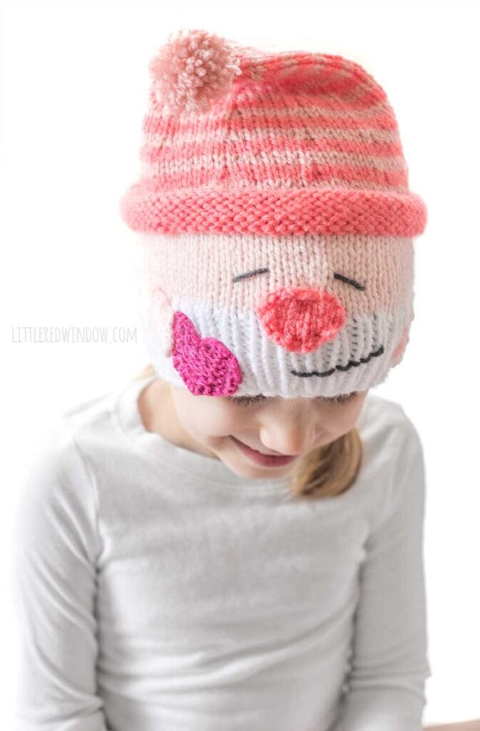 smiling girl in white shirt in front of a white background looking down at her lap wearing a knit hat that looks like a smiling white bearded Valentine's Day gnome holding a hot pink heart in his hand and wearing a stocking cap