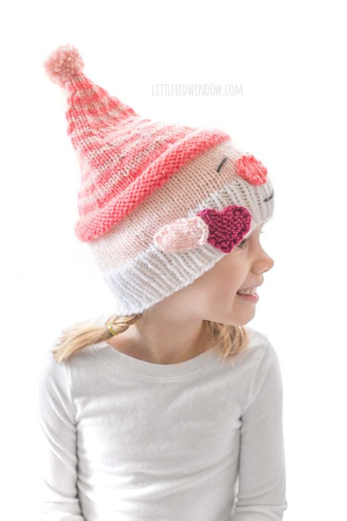 smiling girl in white shirt in front of a white background looking off to the right wearing a knit hat that looks like a smiling white bearded Valentine's Day gnome holding a hot pink heart in his hand and wearing a stocking cap
