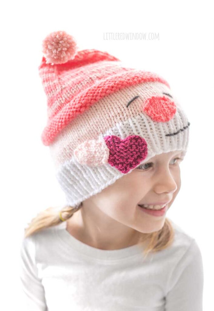 closer view of smiling girl in white shirt in front of a white background looking off to the right wearing a knit hat that looks like a smiling white bearded Valentine's Day gnome holding a hot pink heart in his hand and wearing a stocking cap