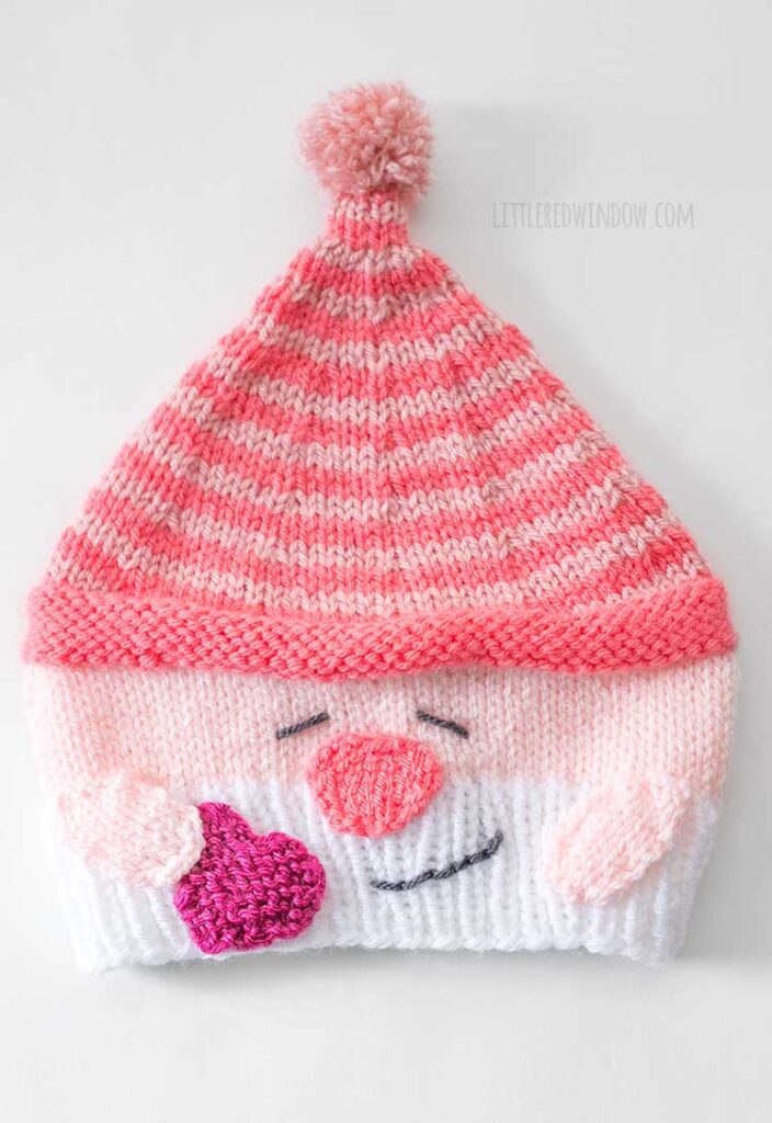 flat lay view of pink and white Valentine Gnome knit hat wiht a white beard smiling face stocking cap holding a hot pink heart in his right hand on a white background