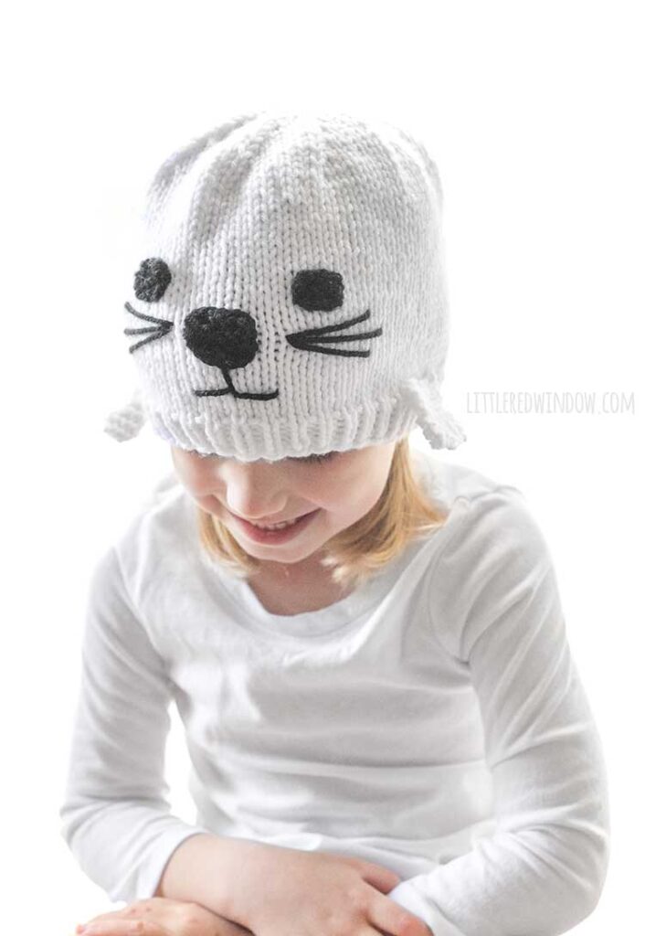 little girl in white shirt and white knit seal hat smiling and looking down and to the left
