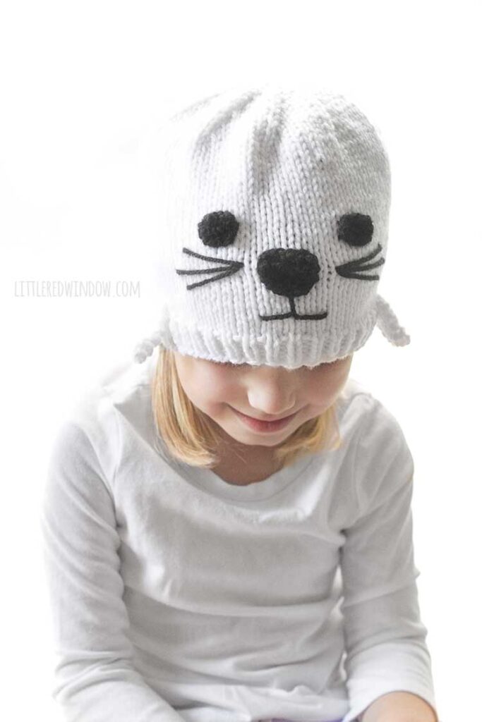 little girl in white shirt and white knit seal hat smiling and looking down and to the right