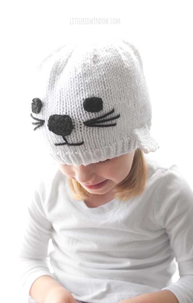 closeup of little girl in white shirt and white knit seal hat smiling and looking down and to the left