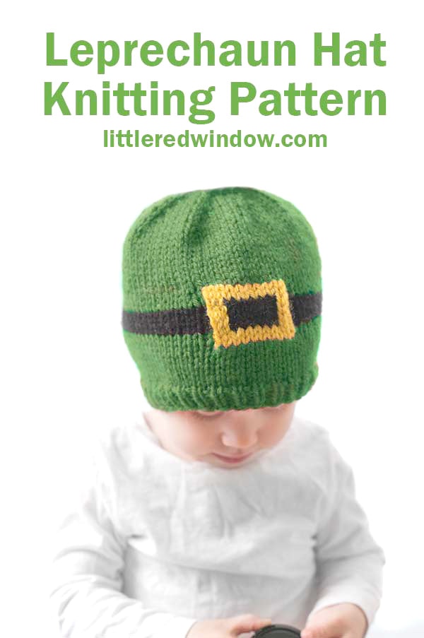 baby in white shirt wearing a green knit hat with a black and gold leprechaun belt around the middle in front of a white background looking down and to the right