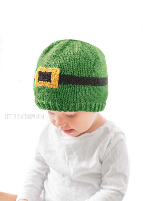 baby in white shirt wearing a green knit hat with a black and gold leprechaun belt around the middle in front of a white background looking down and to the left